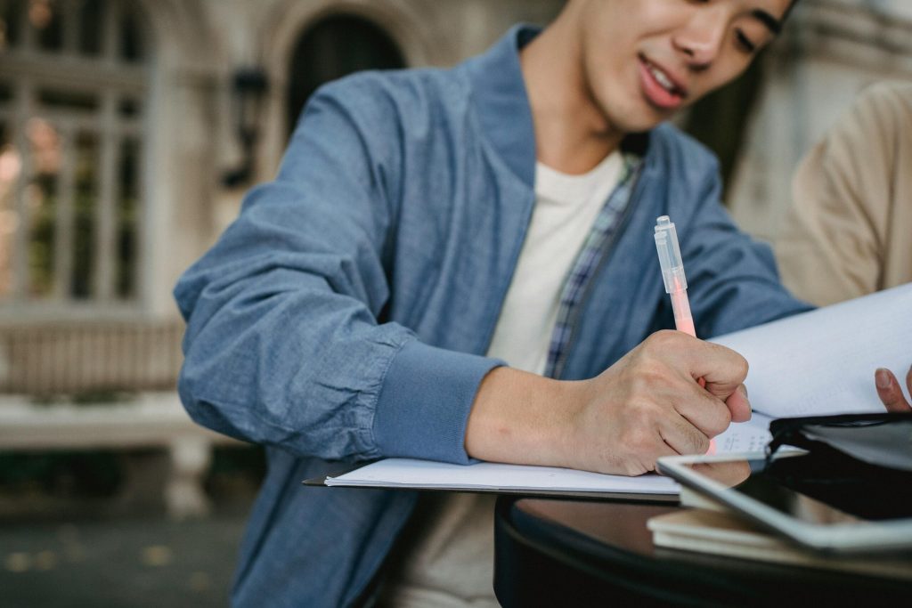 Crop Asian man writing in document while preparing for lesson with friend in park